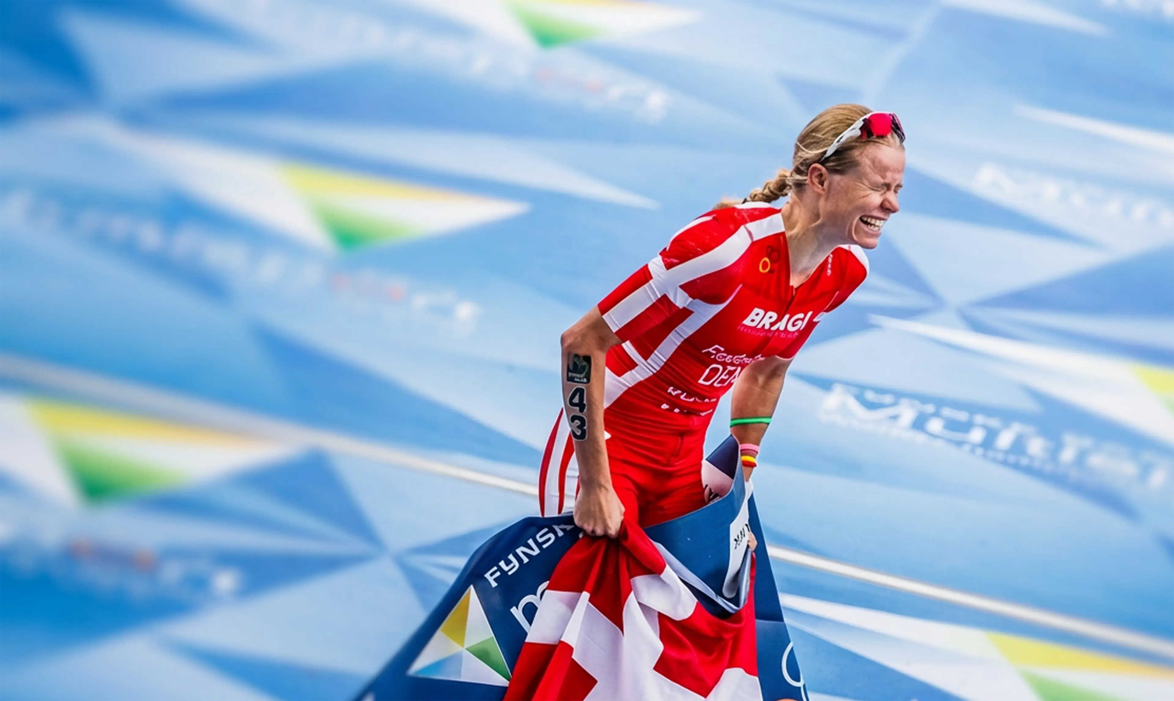 Helle Frederiksen in red and white uniform celebrating victory by holding Denmark flag and smiling on a blue track.