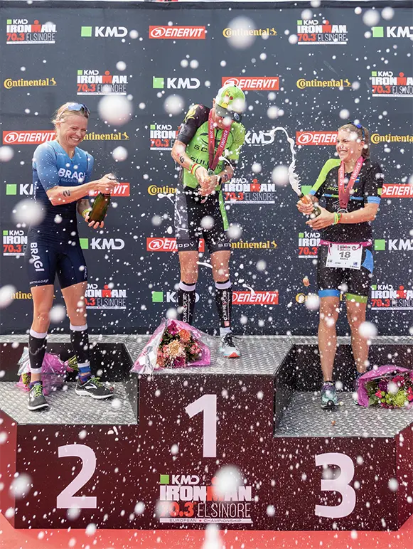 Helle Frederiksen and other athletes on an Ironman 70.3 Elsinore winners podium spraying champagne and celebrating their victory.