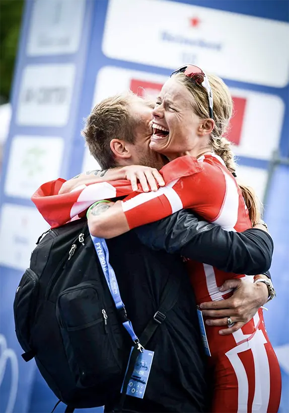 Helle Frederiksen in red and white sportswear joyfully hugging a man with a backpack at an event finish area.