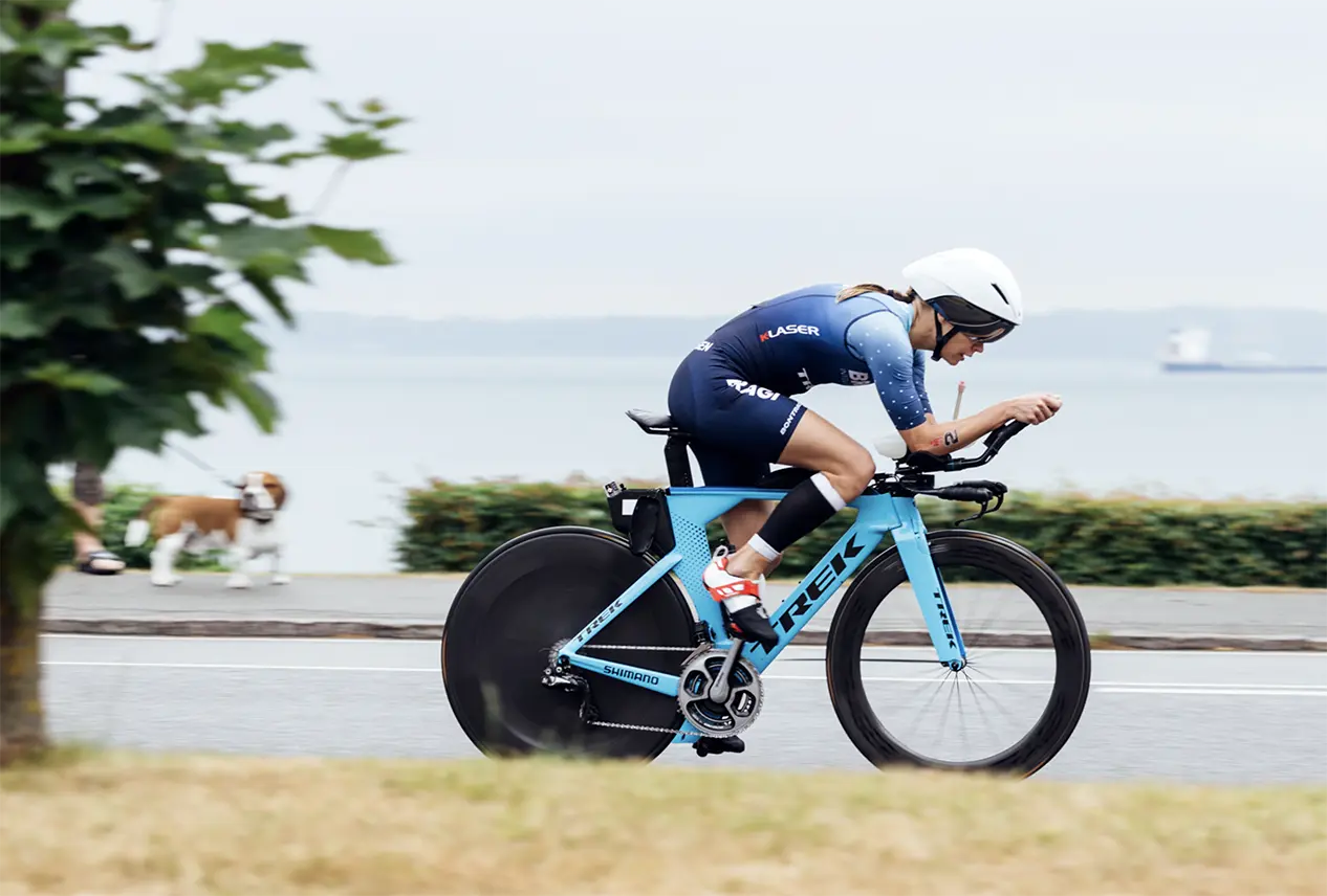 Helle Frederiksen in blue cycling suit and white helmet riding a blue Trek time trial bike on a road near water.