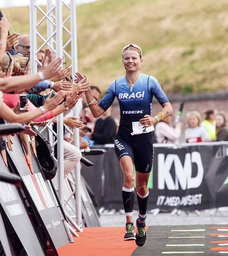 Helle Frederiksen in blue and black BRAGI triathlon suit running and high-fiving spectators at finish line of a race.