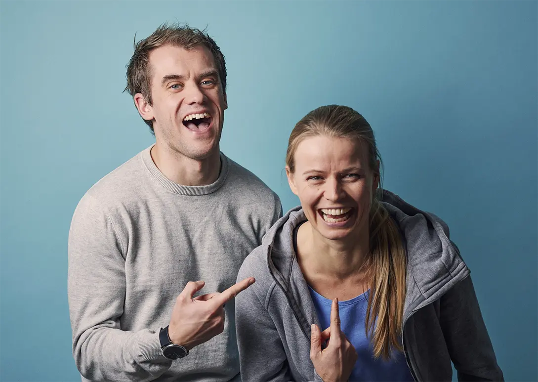 Ben Powell-Frederiksen and Helle Frederiksen smiling and laughing, both pointing fingers at each other against a blue background.
