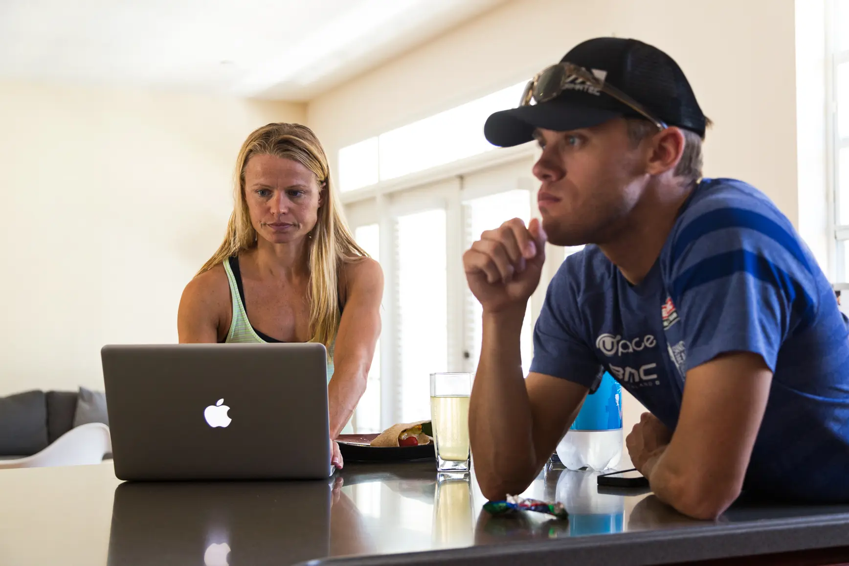 Helle Frederiksen using a laptop on a kitchen counter while Ben Powell-Frederiksen sits beside her, resting his chin on his hand with a glass and a wrapped snack on the counter.
