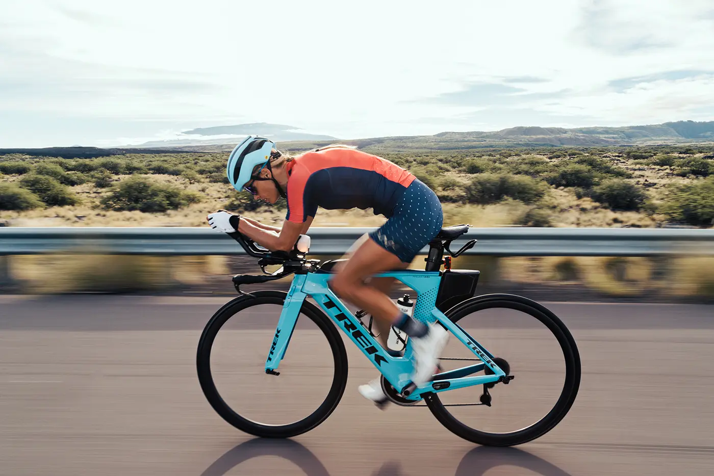 Helle Frederiksen in blue and orange outfit riding a turquoise Trek bike fast on a road with blurred background of vegetation and mountains.