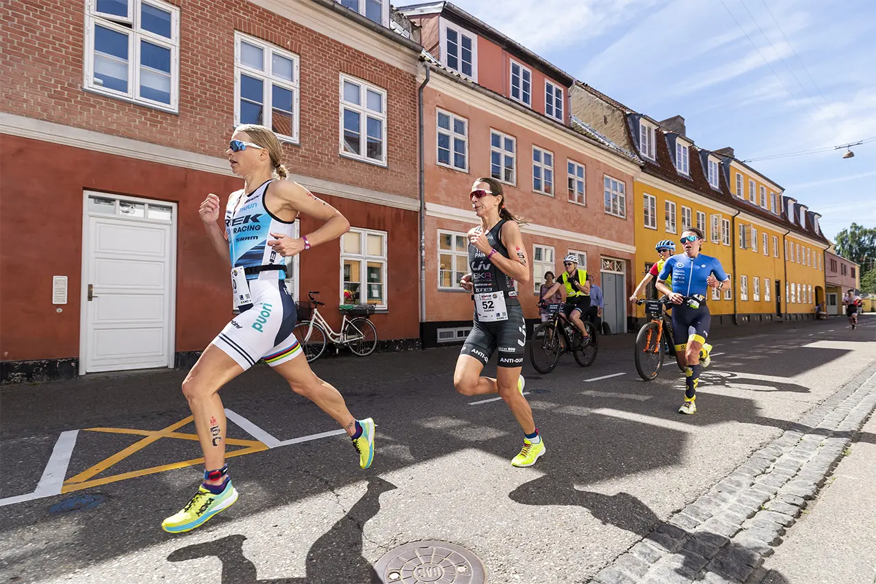 Helle Frederiksen and two female athletes running on a street lined with colorful buildings during a race, followed by two cyclists.