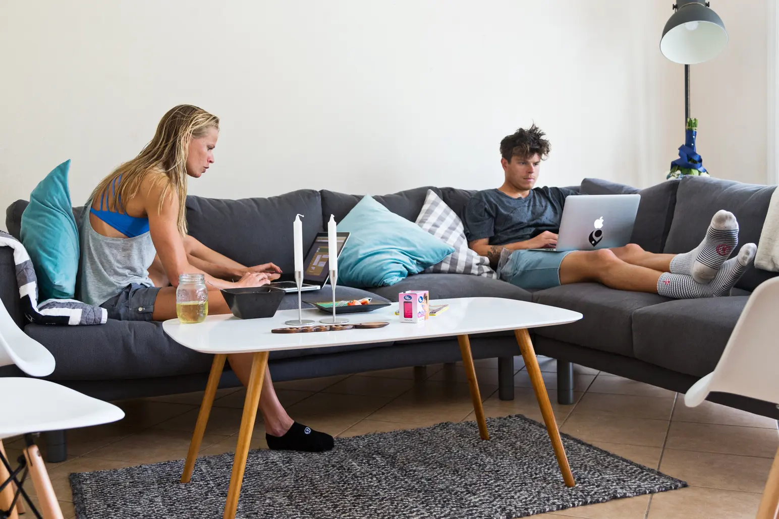 Helle Frederiksen and a man sitting on a gray sectional sofa each working on a laptop with a white coffee table and candles in front of them.