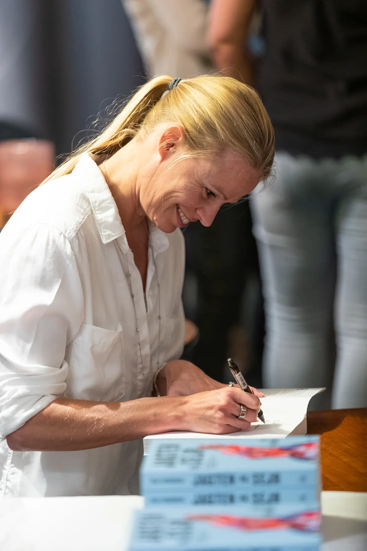 Helle Frederiksen wearing a white shirt signing a book at a table.