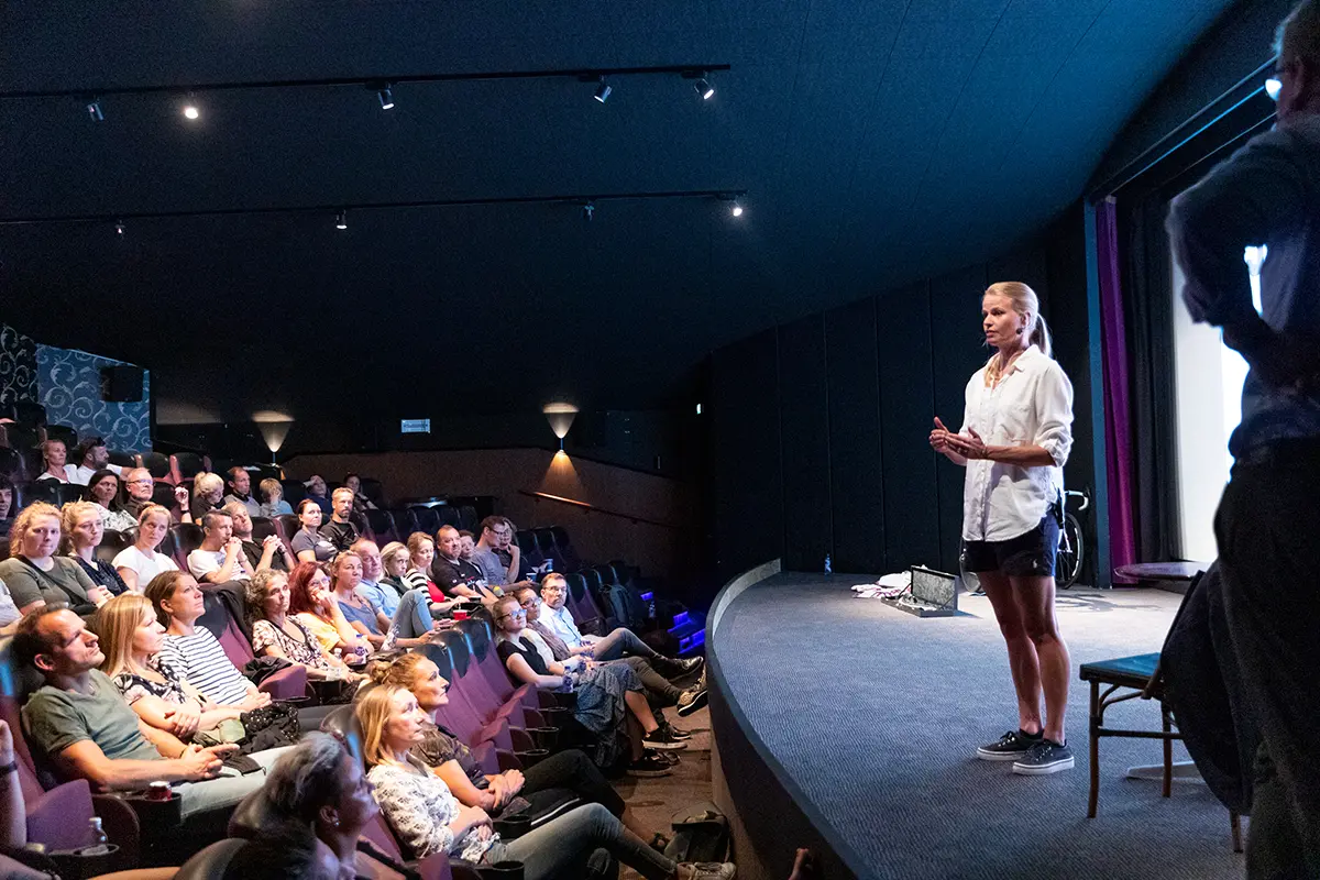 Helle Frederiksen in casual clothing speaking on stage to a seated audience in a dimly lit theater.