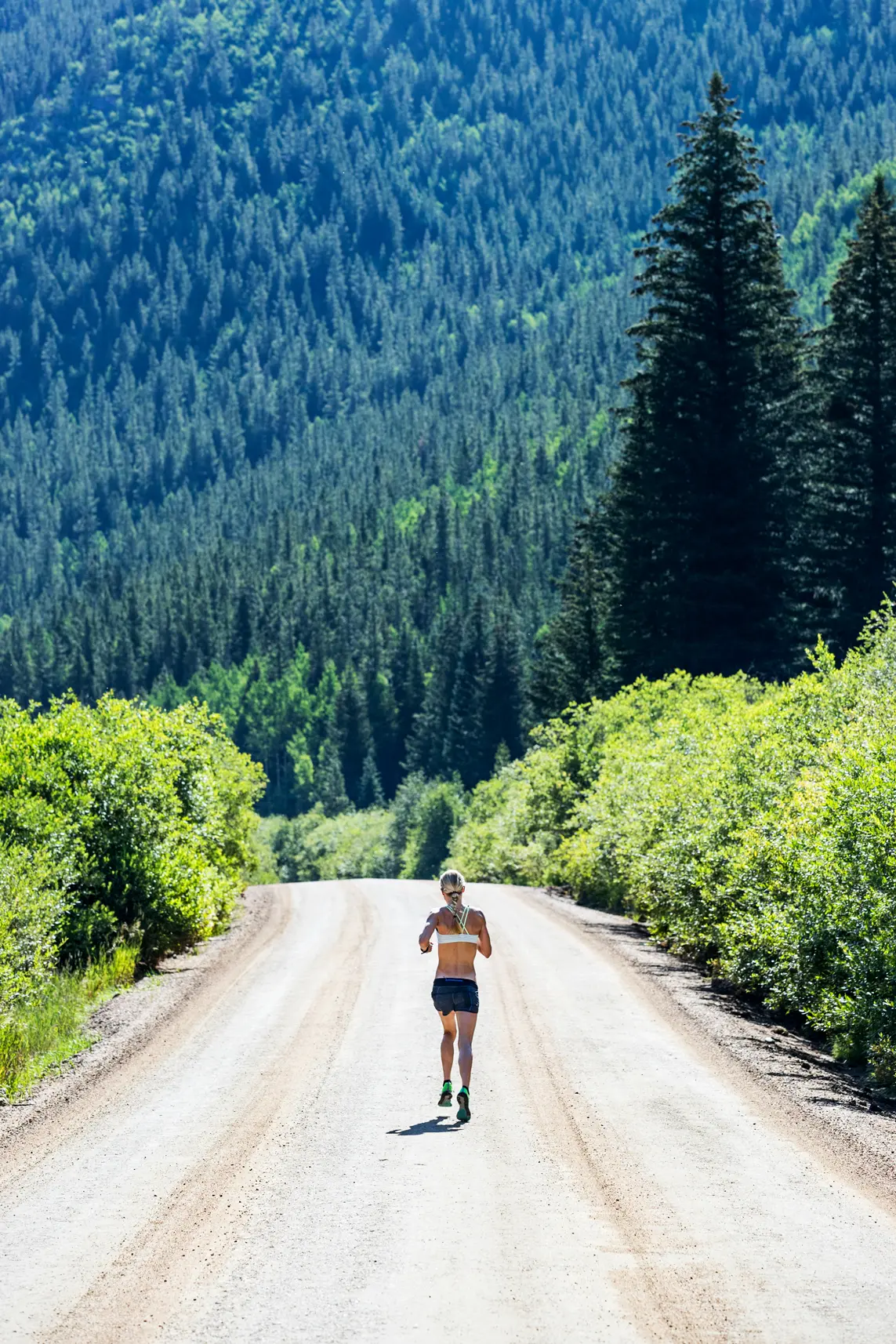 Helle Frederiksen jogging on a wide dirt road surrounded by green bushes and tall pine trees in a forested mountain area.