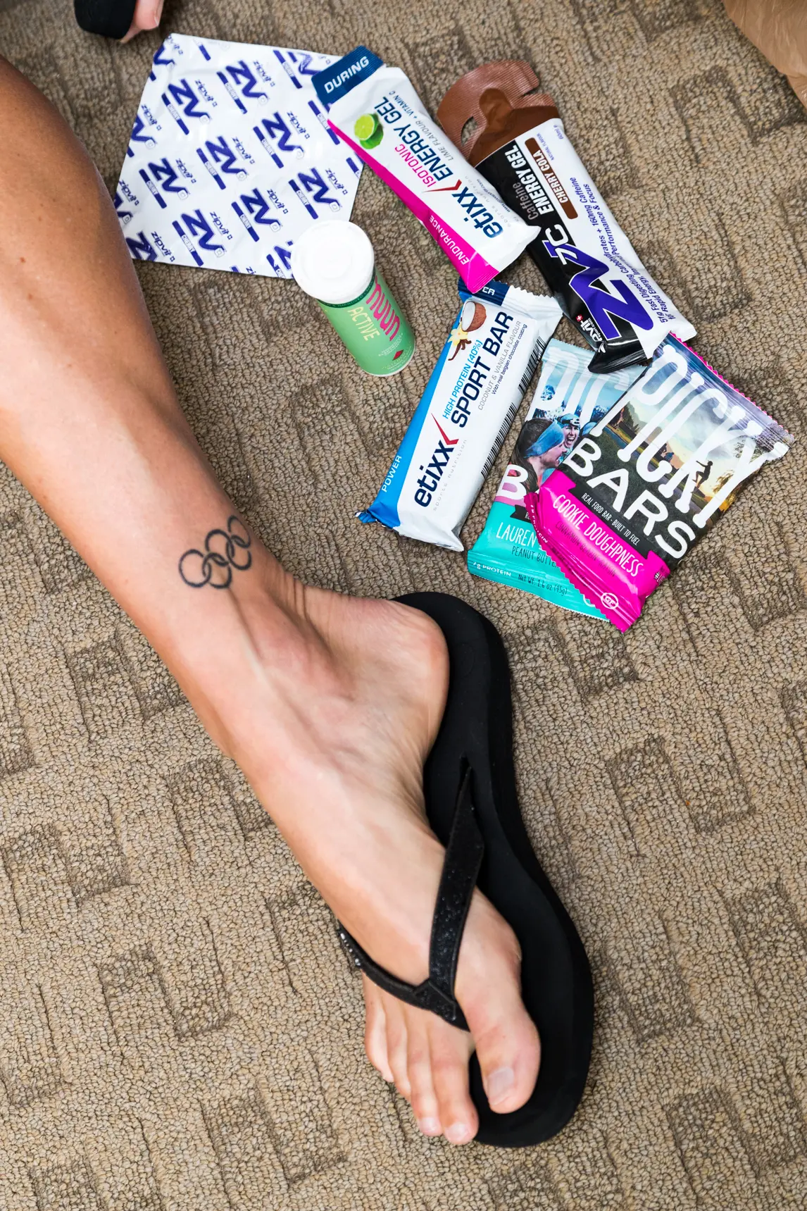 Helle Frederiksen's foot wearing a black flip-flop with an Olympic rings tattoo on the ankle surrounded by various energy gels and nutrition bars on a carpeted floor.