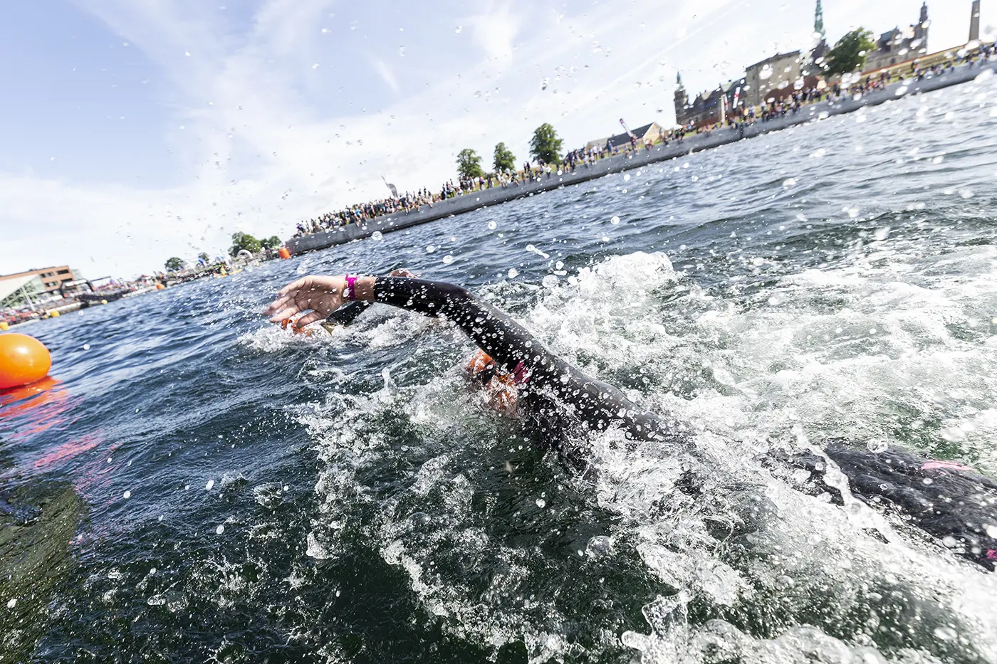 Helle Frederiksen in a wetsuit and orange swim cap swimming freestyle in open water near a shore crowded with spectators under a blue sky.