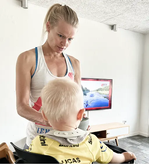 Helle Frederiksen wearing a sleeveless top interacts with a small child with blonde hair seated in a high chair in a modern, minimalist room.
