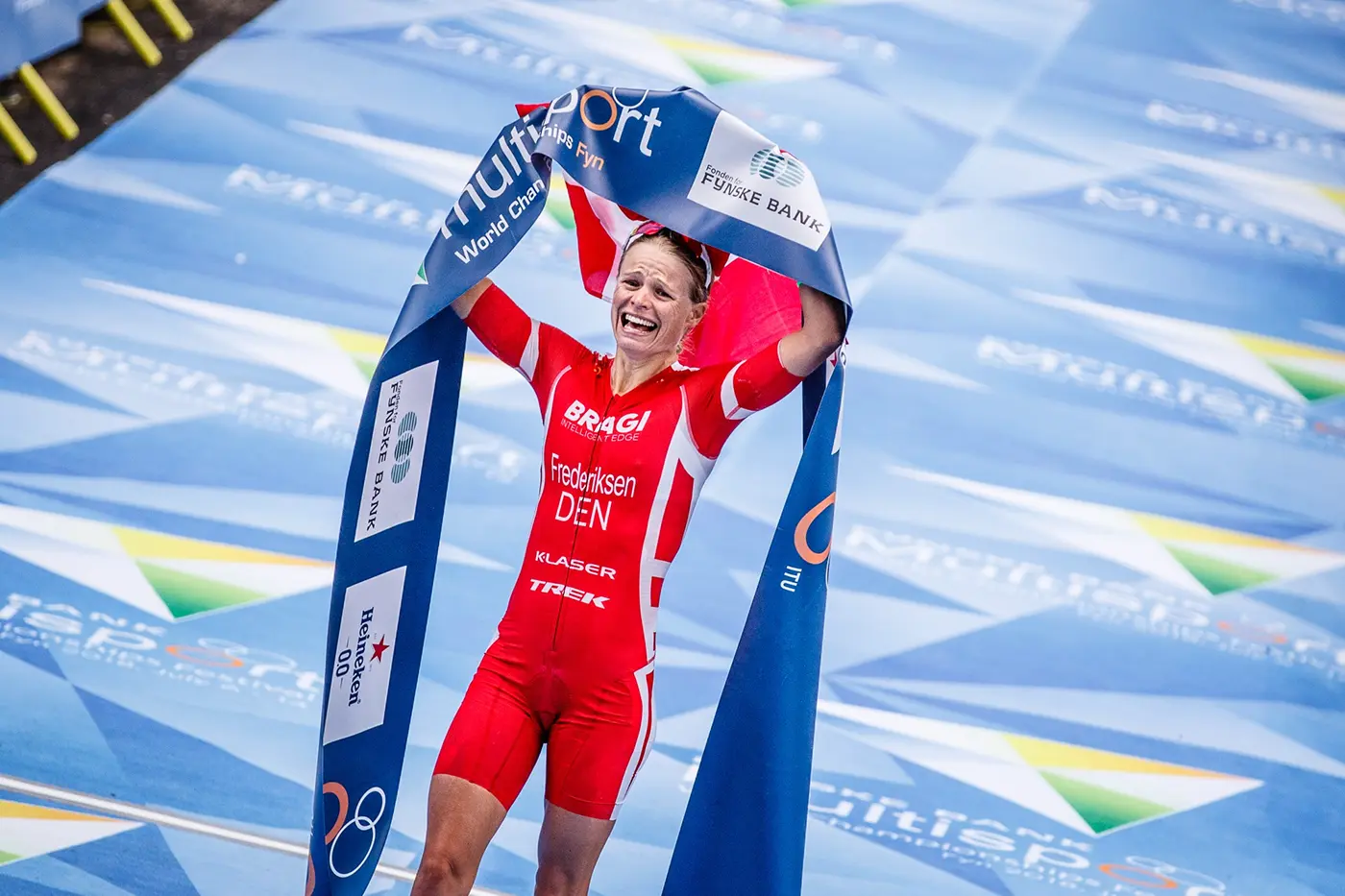 Helle Frederiksen in red triathlon suit celebrating victory by lifting a blue banner at the finish line of a race event.