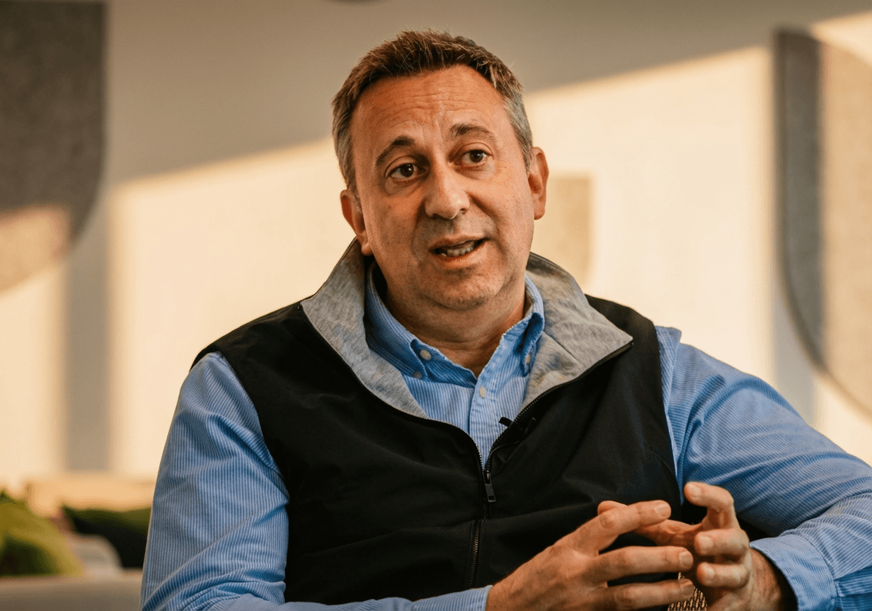 Middle-aged man in a blue shirt and black vest speaking with hand gestures in a softly lit room.