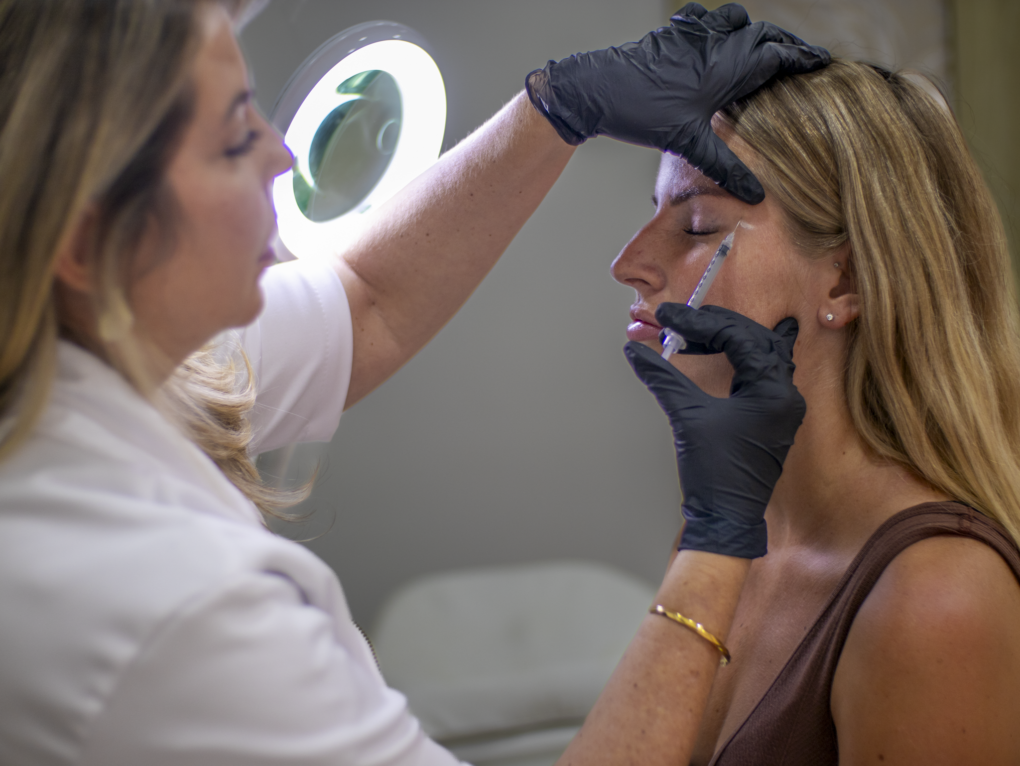 Woman with eyes closed receiving eyebrow injection from professional wearing black gloves and white coat.
