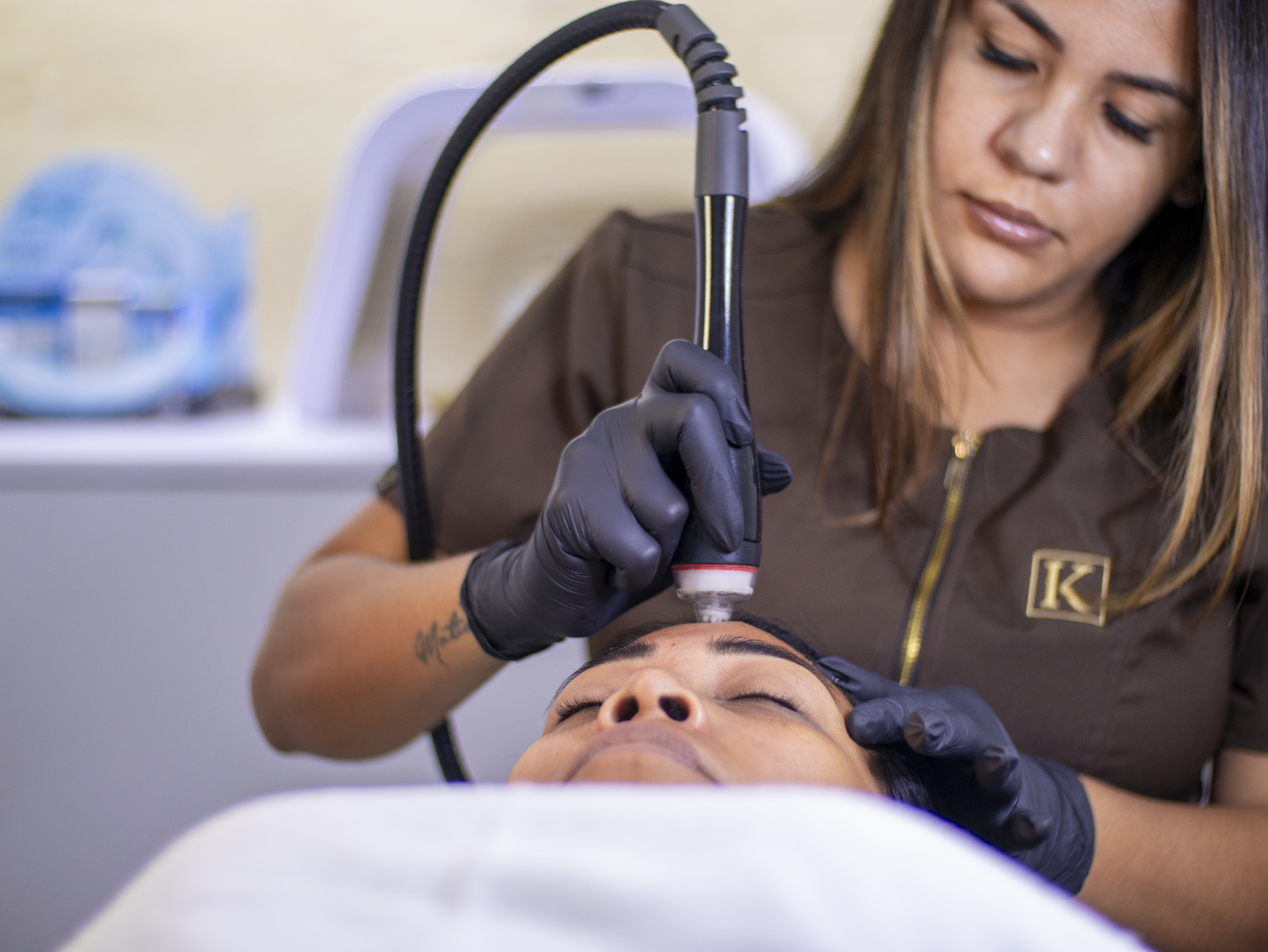 Woman receiving facial treatment with a handheld skincare device by a technician wearing black gloves.