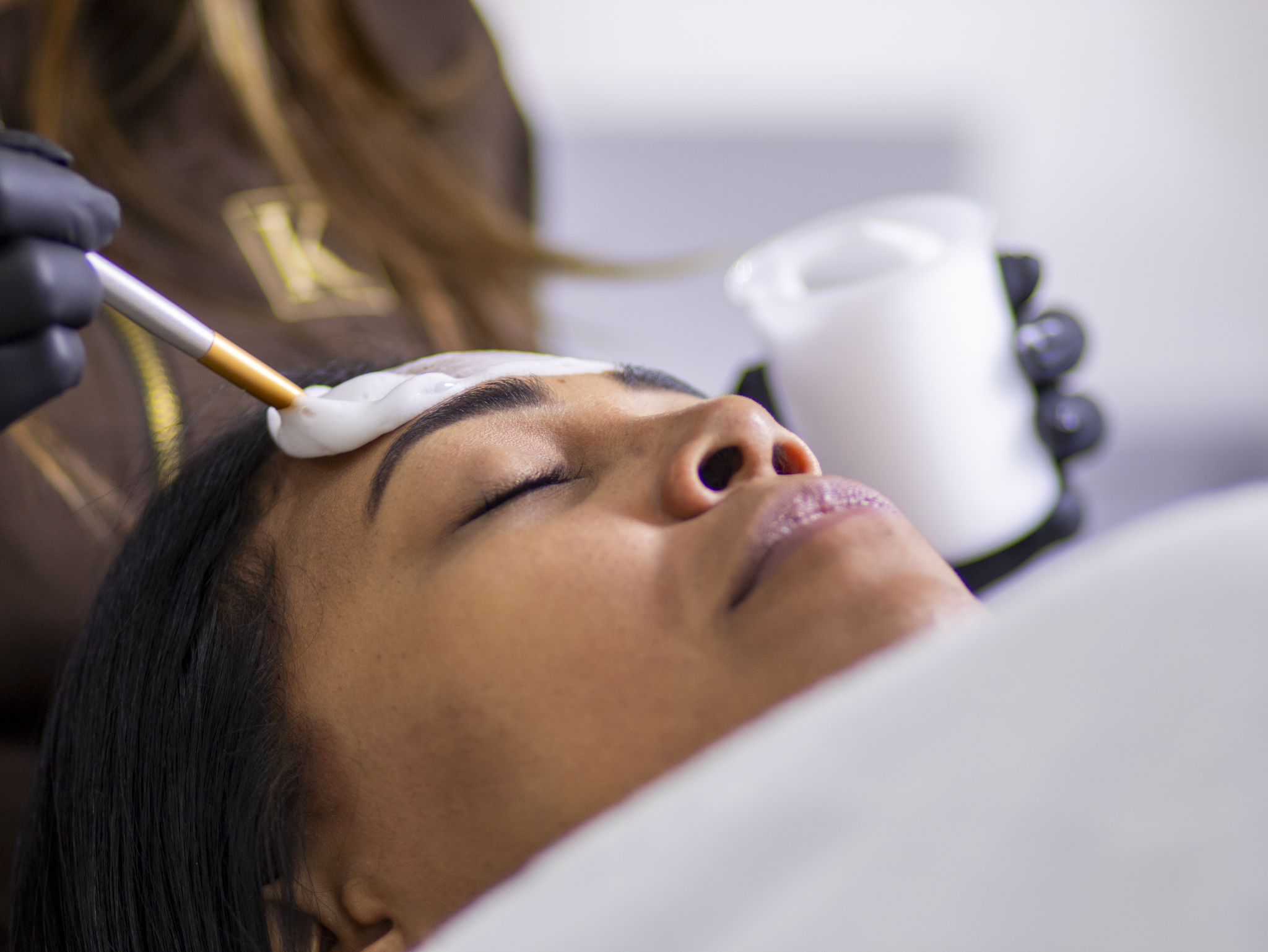 Woman lying down with eyes closed while a cosmetic cream is applied on her forehead with a brush.
