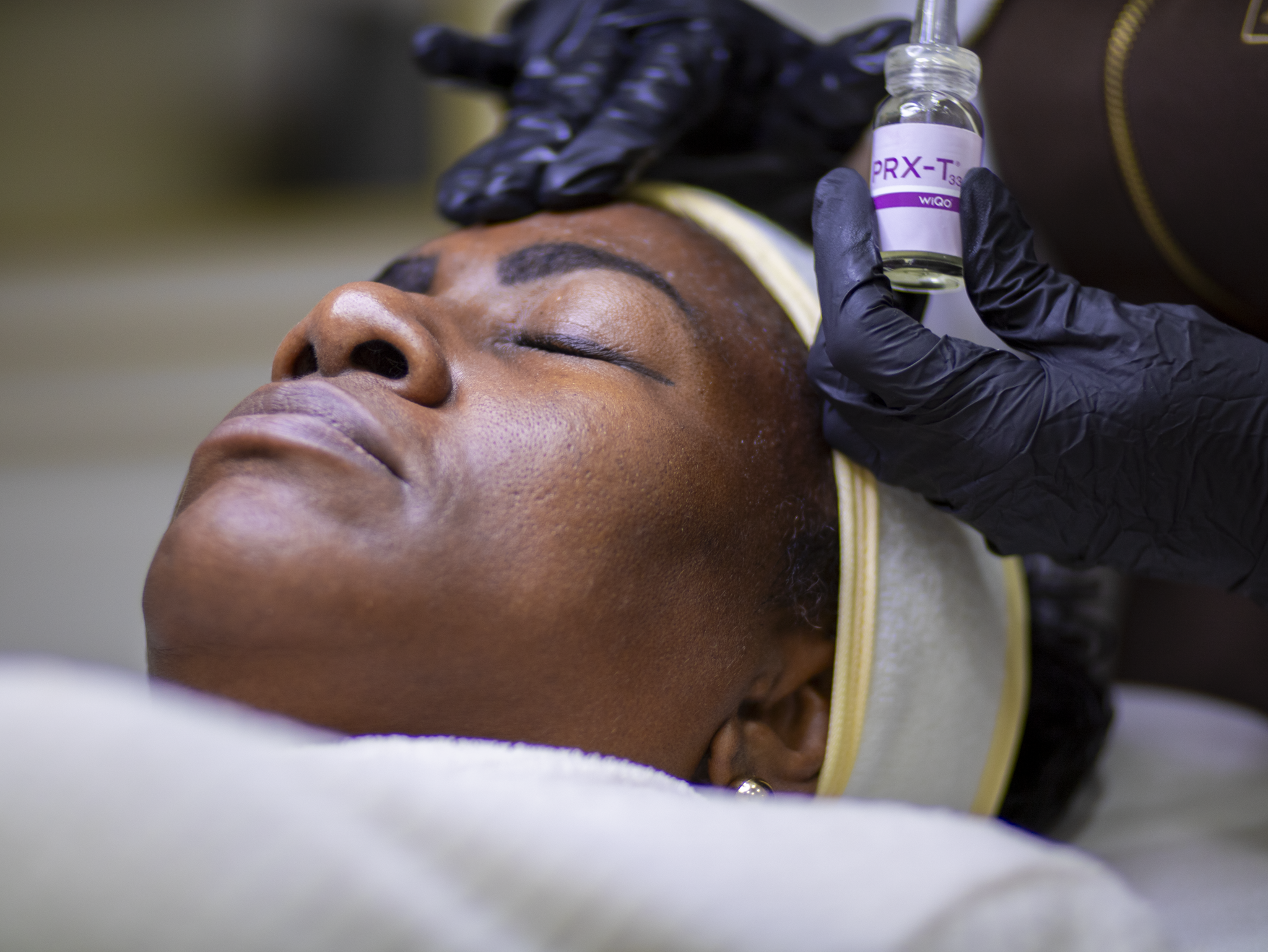 Woman lying down with eyes closed receiving a facial mask treatment applied with a brush on her forehead and cheeks.