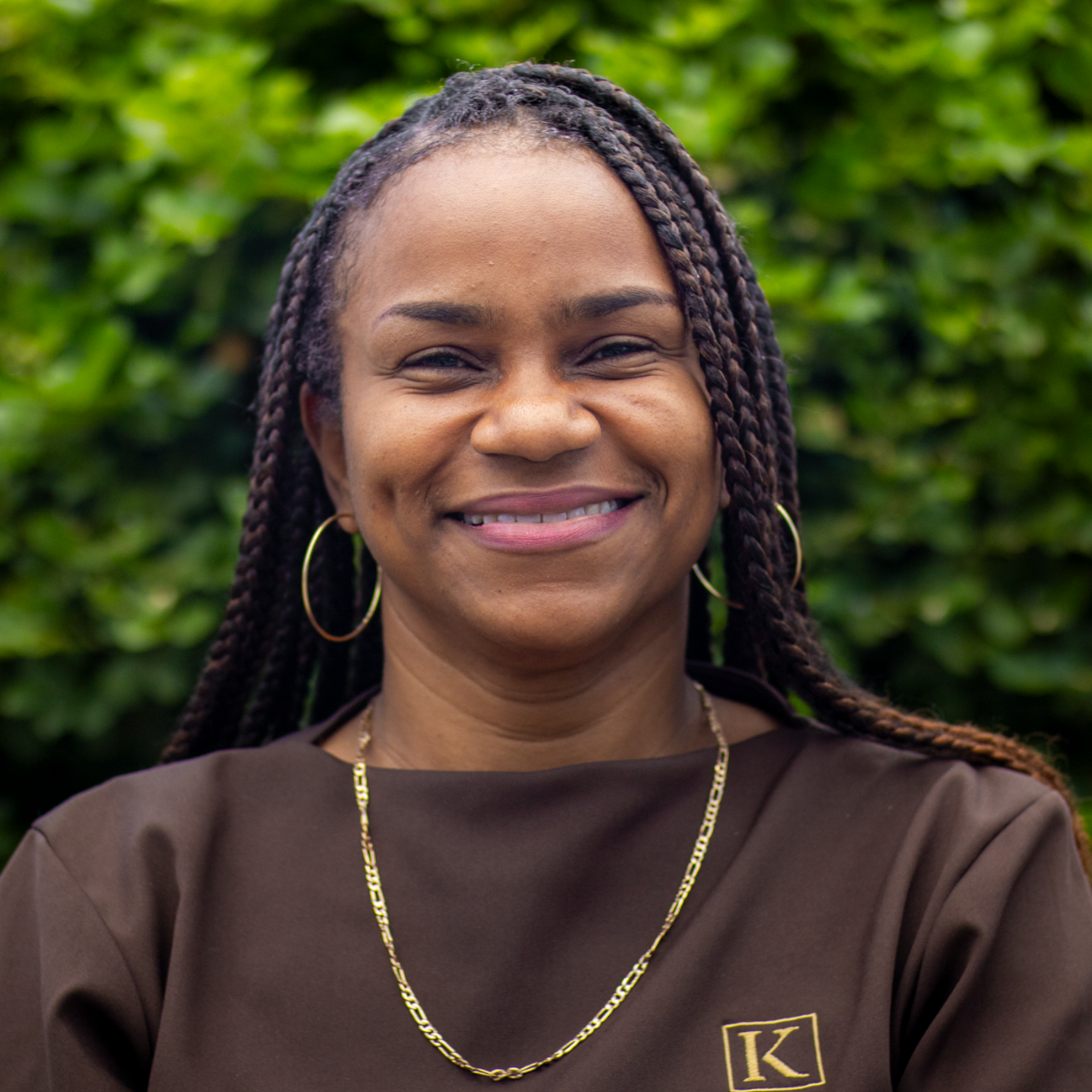 Smiling woman with braided hair, gold hoop earrings, and gold chain necklace in front of green foliage.