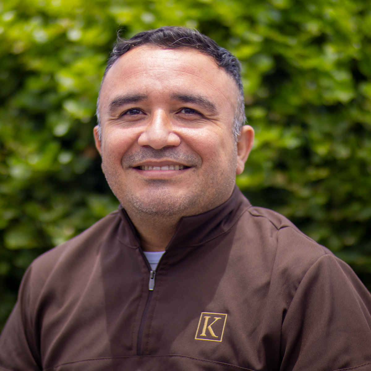 Smiling man wearing a brown jacket with a K logo, standing outdoors with green leaves background.
