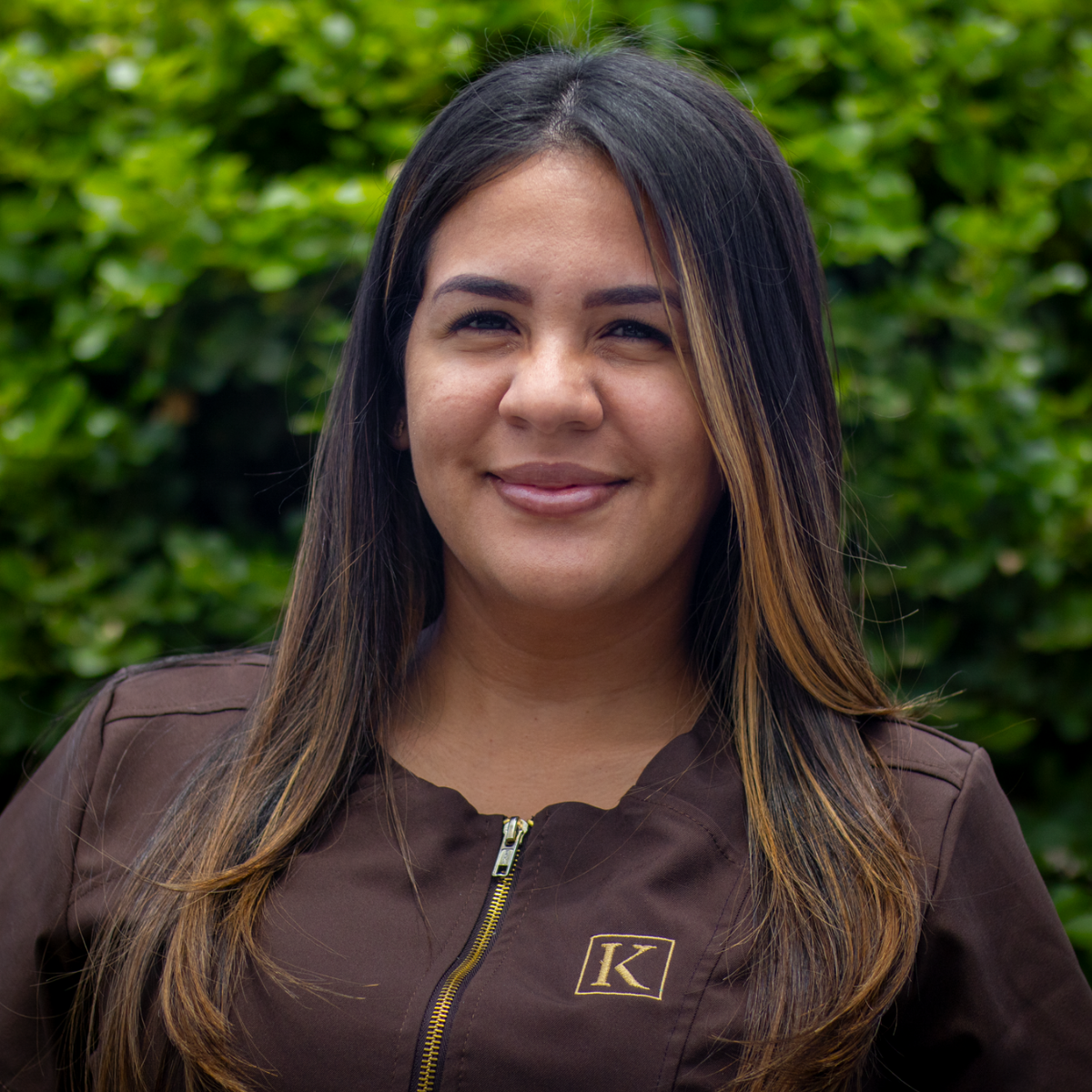 Smiling woman with long hair wearing a brown zip-up jacket with a gold 'K' logo, outdoors.