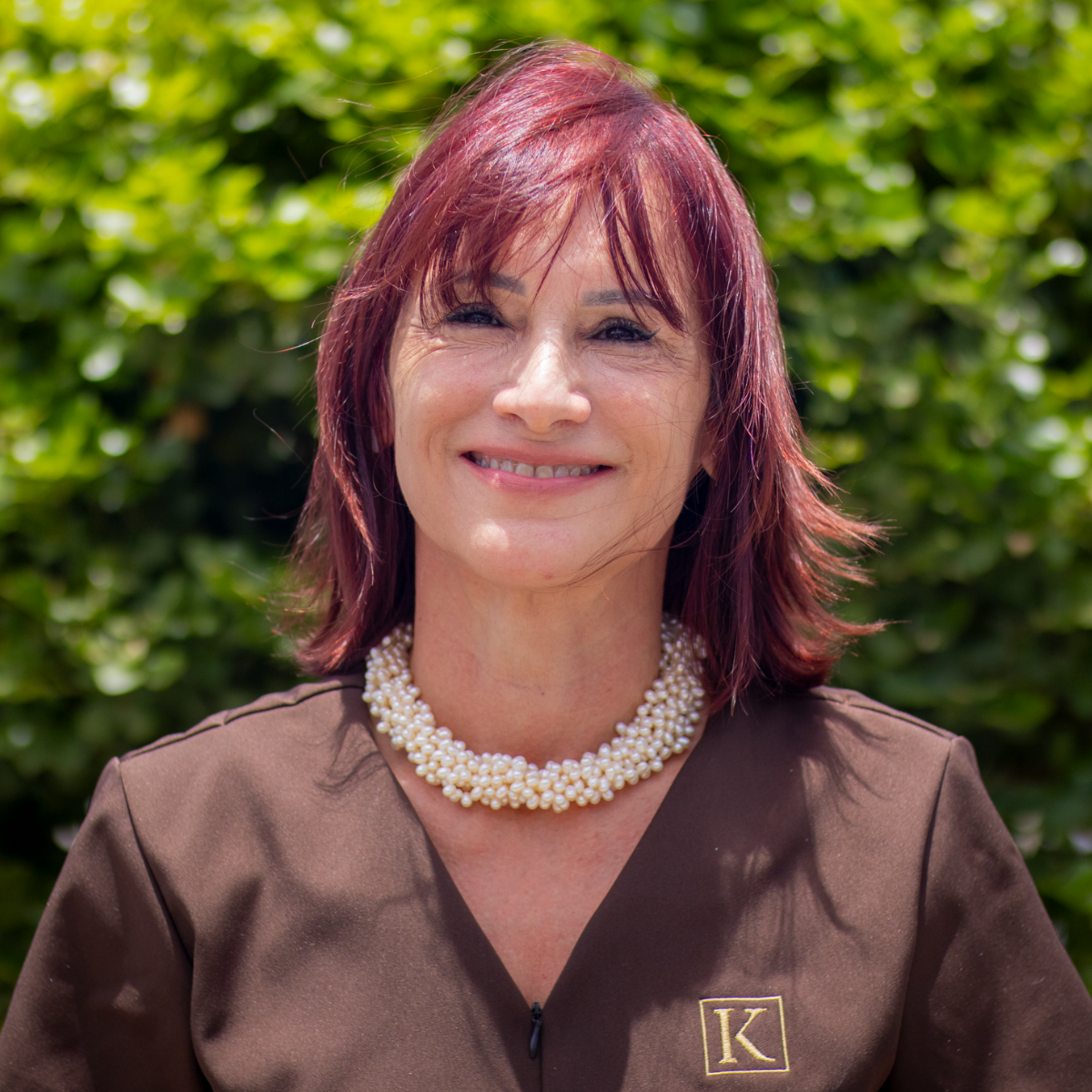 Smiling woman with reddish hair wearing a brown top and pearl necklace, standing outdoors with greenery.