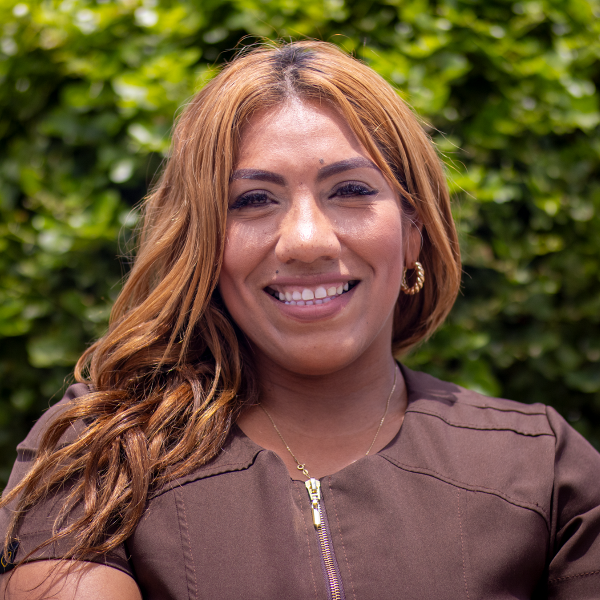 Smiling woman with long light brown hair wearing a brown top and gold hoop earrings outdoors.
