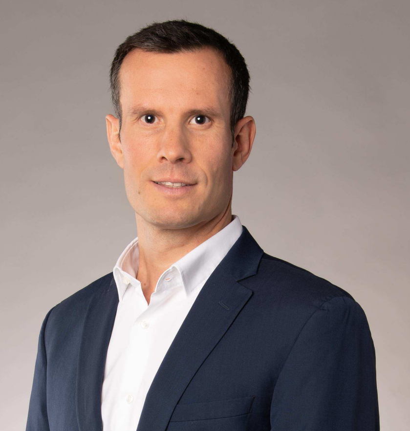 Man wearing a dark suit jacket and white shirt against a plain gray background.