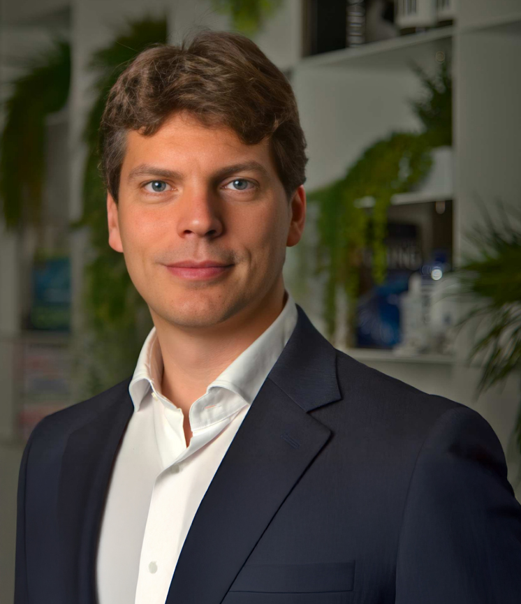 Portrait of a man in a dark suit and white shirt with a blurred office background with plants.