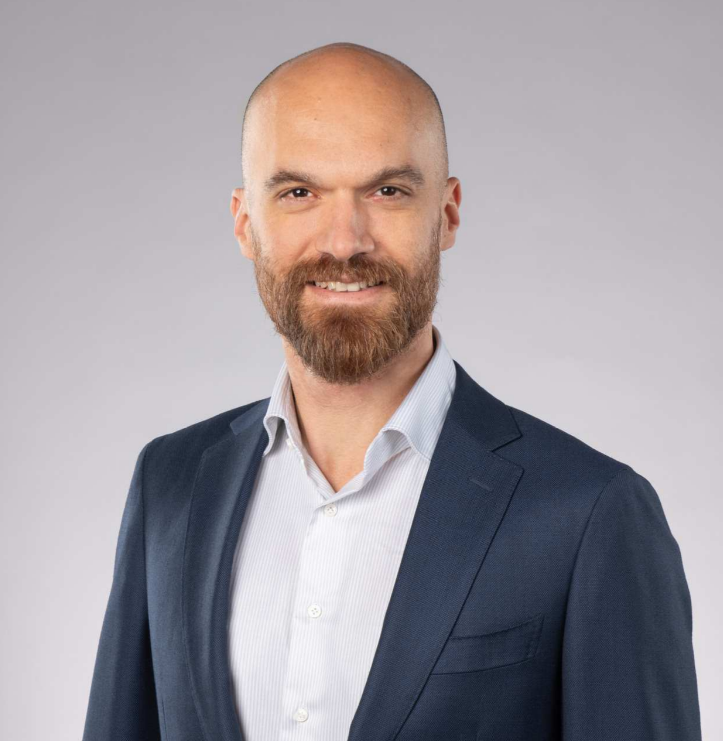 Bald man with beard wearing a navy blazer and white shirt, smiling against a gray background.