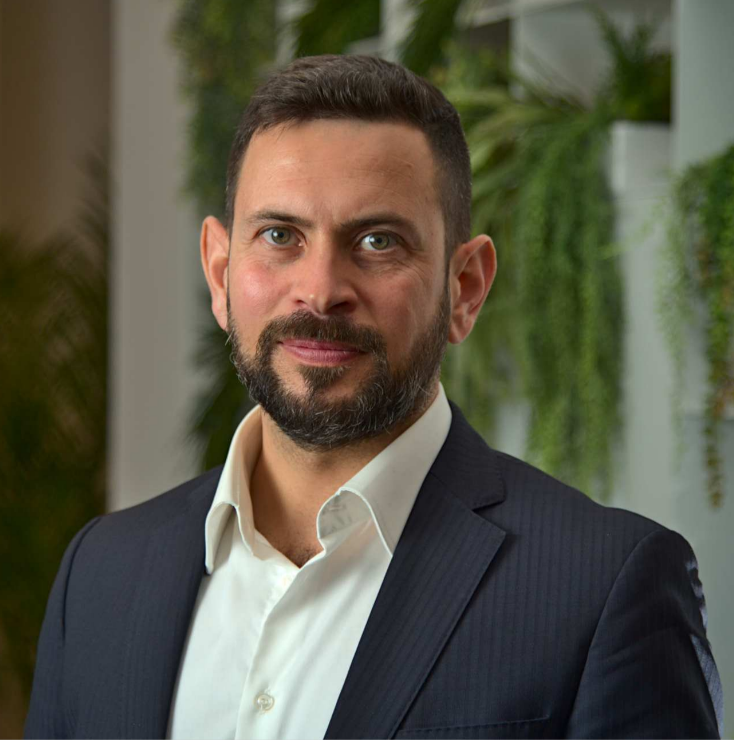Portrait of a man with beard wearing a dark suit and white shirt, with blurred green plants in the background.