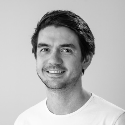 Smiling man with short dark hair wearing a white shirt against a plain light background.
