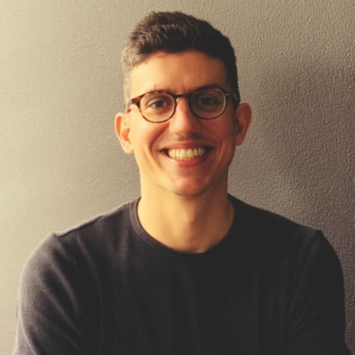 Smiling man with short hair and glasses wearing a dark sweater against a plain wall.
