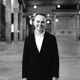 Smiling man in a black suit and white shirt standing in a large industrial space.