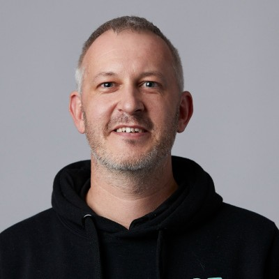 Smiling middle-aged man with short gray hair and beard wearing a black hoodie against a gray background.