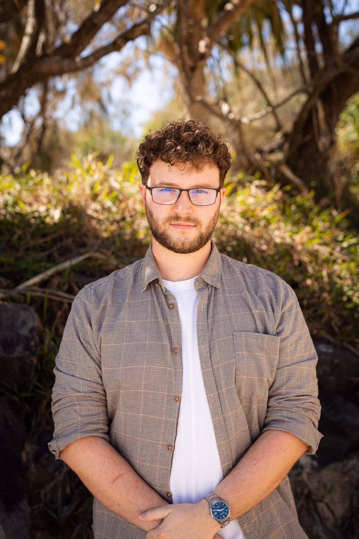 Young man with curly hair and glasses wearing a plaid shirt and white t-shirt standing outdoors with trees and greenery in the background.