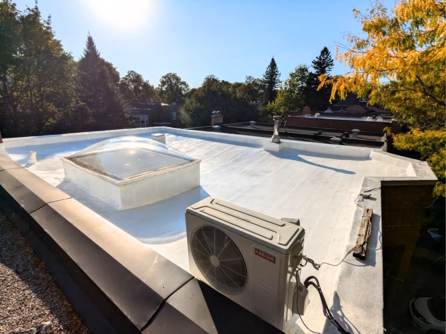 Flat white rooftop with a square skylight dome and an outdoor air conditioning unit, surrounded by trees under a clear blue sky.