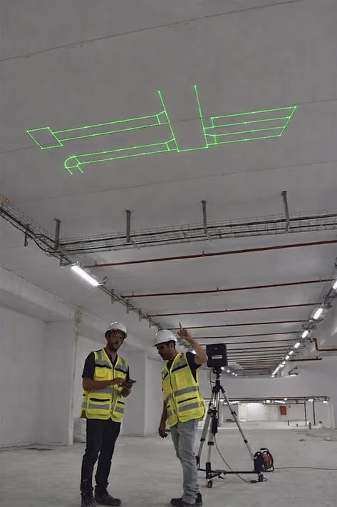 Two construction workers using laser projection to display BIM model ductwork geometry on a ceiling, showing exact HVAC layout positions during field installation