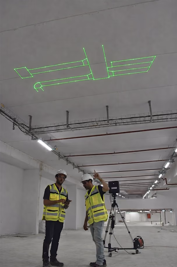 Two MEP workers using laser layout projection to mark HVAC ductwork geometry from a BIM model directly onto a concrete ceiling during mechanical layout