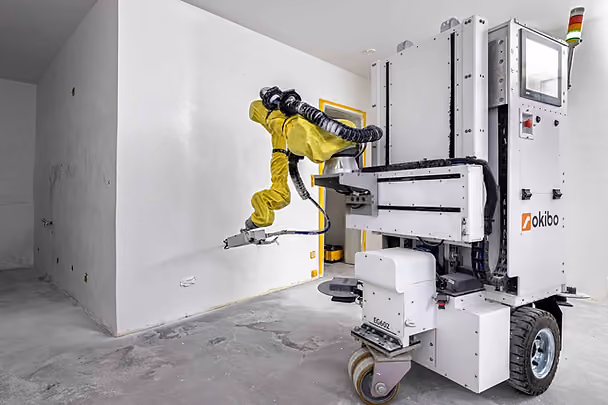 Robot with yellow arm painting a white wall in a plain room. Machine labeled "okibo" and "EG602" with gray floor and walls.
