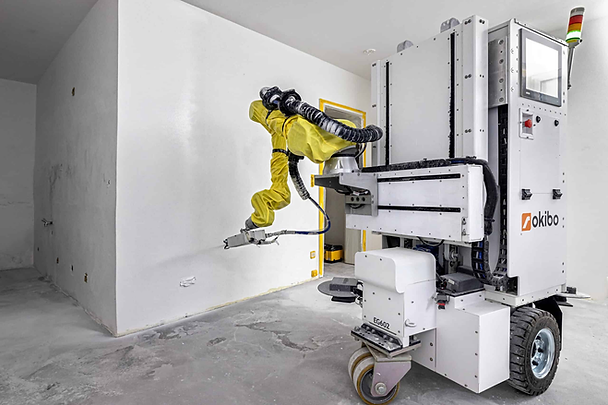 Robot with yellow arm painting a white wall in a plain room. Machine labeled "okibo" and "EG602" with gray floor and walls.