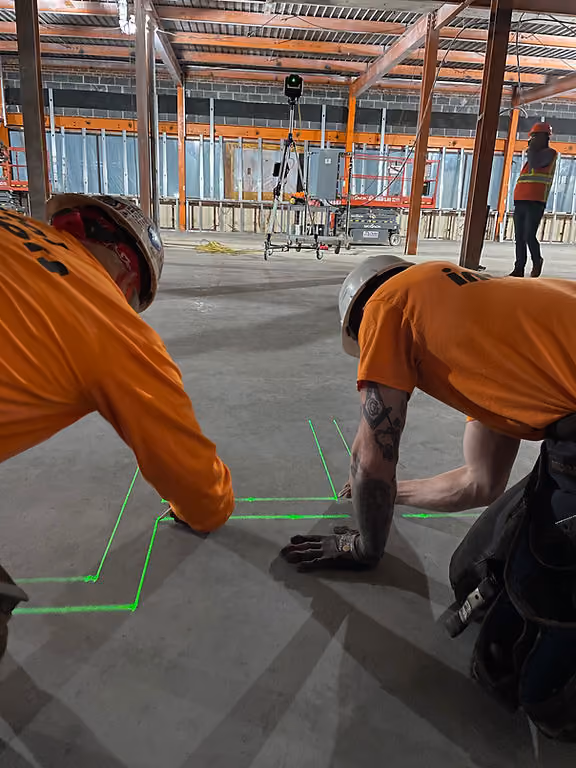 Construction workers in orange shirts and helmets mark a concrete floor with green laser lines. Industrial site with scaffolding and laser tool.