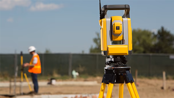 Surveying equipment in focus on a construction site, with a worker in an orange vest and white helmet in the blurred background. Sunny day.