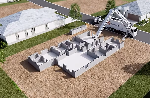 A truck with "Hadrian X" builds a concrete house foundation on a grassy lot. Nearby are trees, a road, and houses with grey roofs.