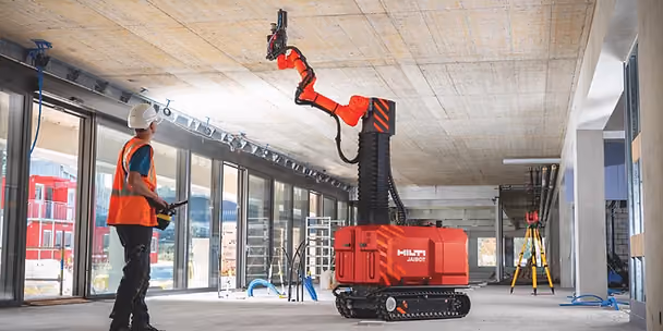 Construction worker observes a red Hilti Jaibot drilling into a concrete ceiling inside a building under construction, with large windows.