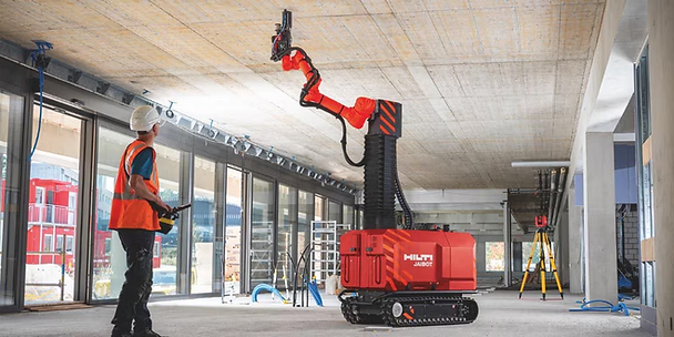 Construction worker observes a red Hilti Jaibot drilling into a concrete ceiling inside a building under construction, with large windows.