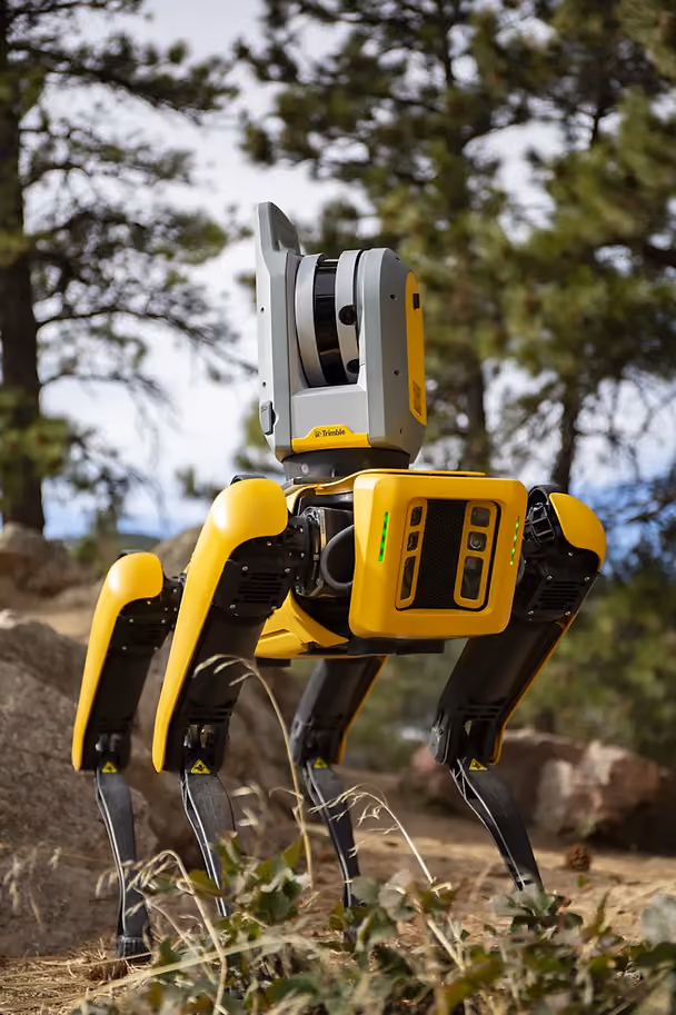 Yellow and gray robotic dog with Trimble logo walks in a forest setting. Pine trees and rocks in the background, creating a tech-nature mood.