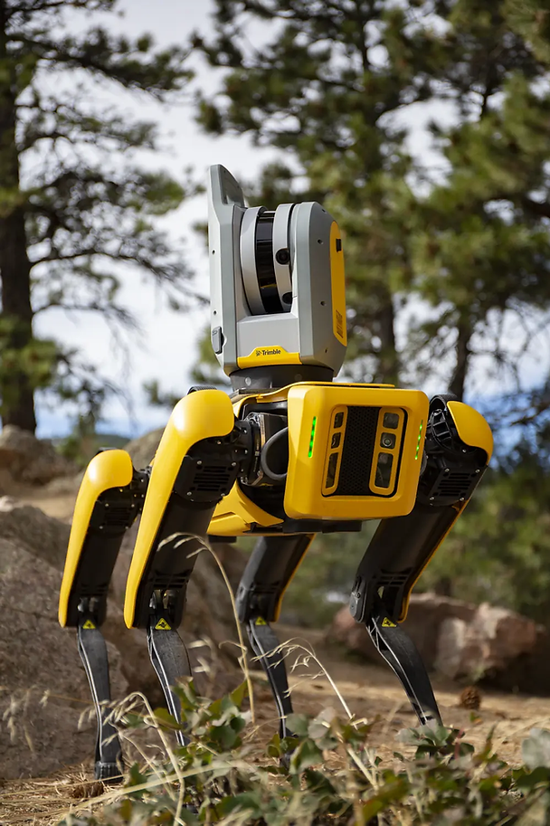 Yellow and gray robotic dog with Trimble logo walks in a forest setting. Pine trees and rocks in the background, creating a tech-nature mood.