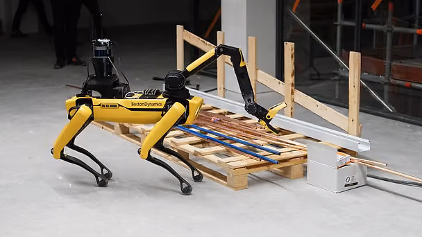 A yellow Boston Dynamics robot with an arm examines construction materials on a wooden pallet in an industrial setting.