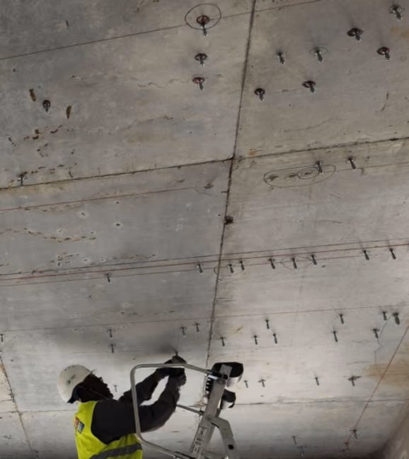 MEP worker installing mechanical hanger anchors into a concrete ceiling during field layout, with multiple pre-drilled hanger points visible across the slab surface